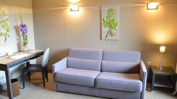 A room with a grey sofa, a desk with a chair, a small table with a lamp, and wall-mounted lights.