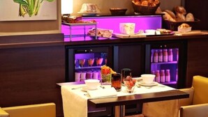 A breakfast buffet with various food items and beverages, a seating area with chairs, and a refrigerator with drinks. - Hôtel les Rives (Guebwiller)