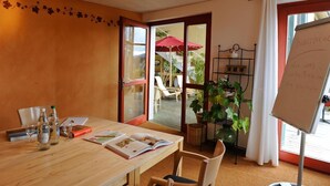A room with a wooden table, books, and a whiteboard. There is a door leading to an outdoor area with a red umbrella and chairs. - EcoHotel L'Aubier (Rochefort)
