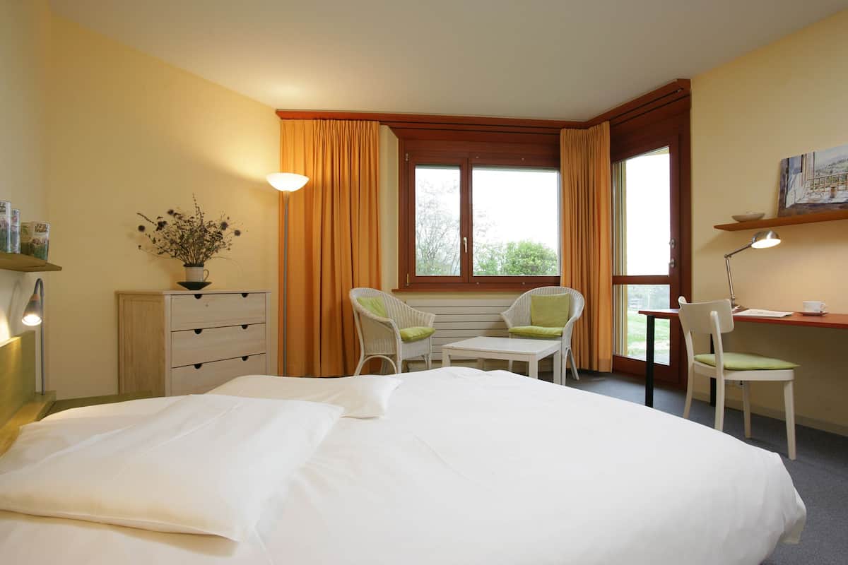 A hotel room with a large bed, a desk with two chairs, a window with curtains, a small table, and a wall-mounted shelf with a vase of flowers.