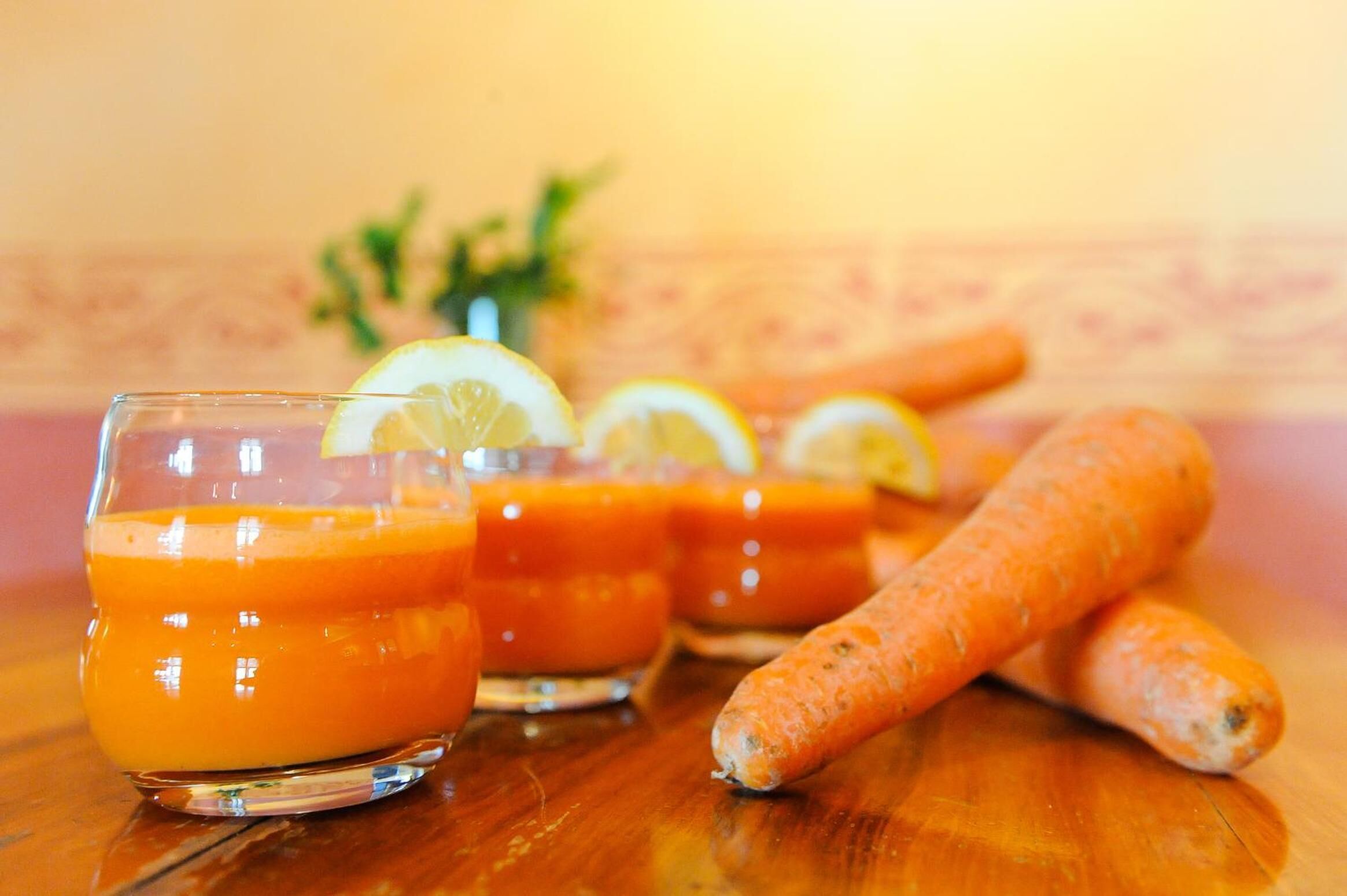 Three glasses of orange juice with lemon slices, a bunch of carrots, and a plant in the background.