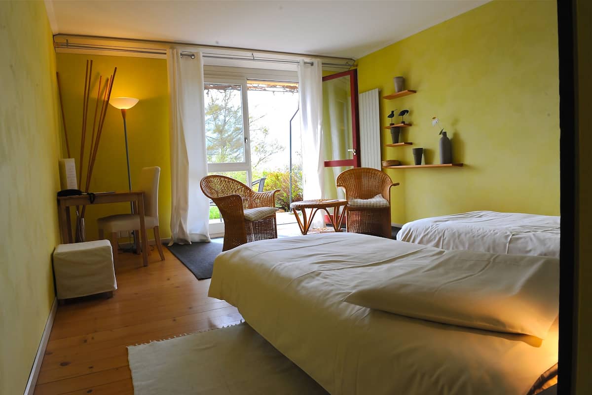 A room with a bed, two wicker chairs, a small table, a lamp, and a shelf with decorative items.