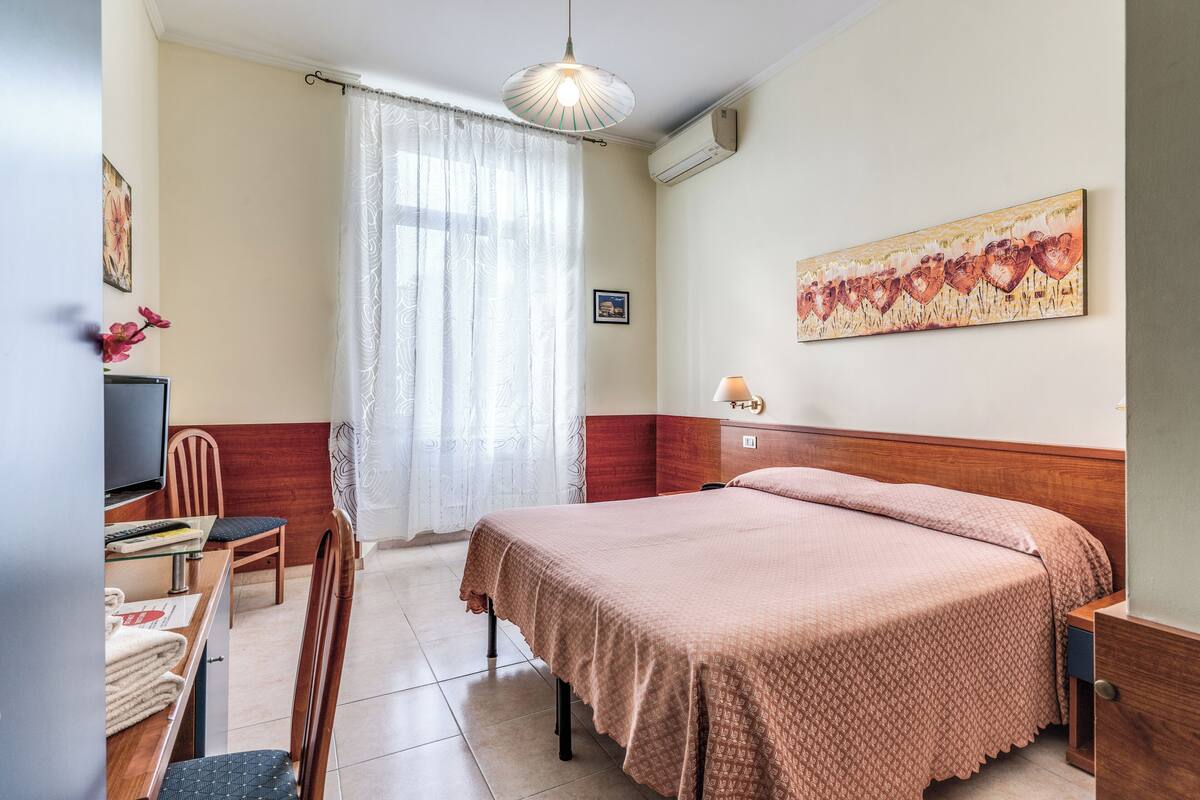 A hotel room with a bed, a desk with a chair, a television, and a window with curtains.