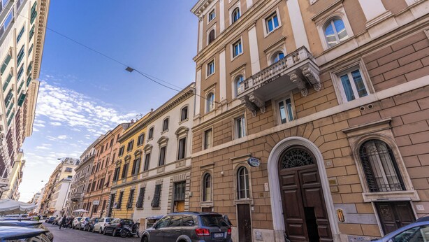 Fila di auto parcheggiate lungo una strada, con un edificio che presenta un ampio ingresso ad arco e un cartello che indica un albergo.