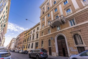 Fila de coches aparcados a lo largo de una calle, con un edificio que tiene una entrada abovedada grande y un cartel que indica un hotel.