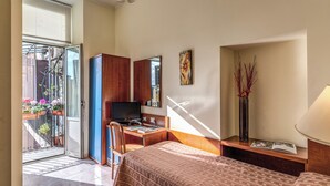Camera d'albergo con un letto, una scrivania con un computer, una sedia, una TV, una finestra con persiane e un balcone con piante in vaso.