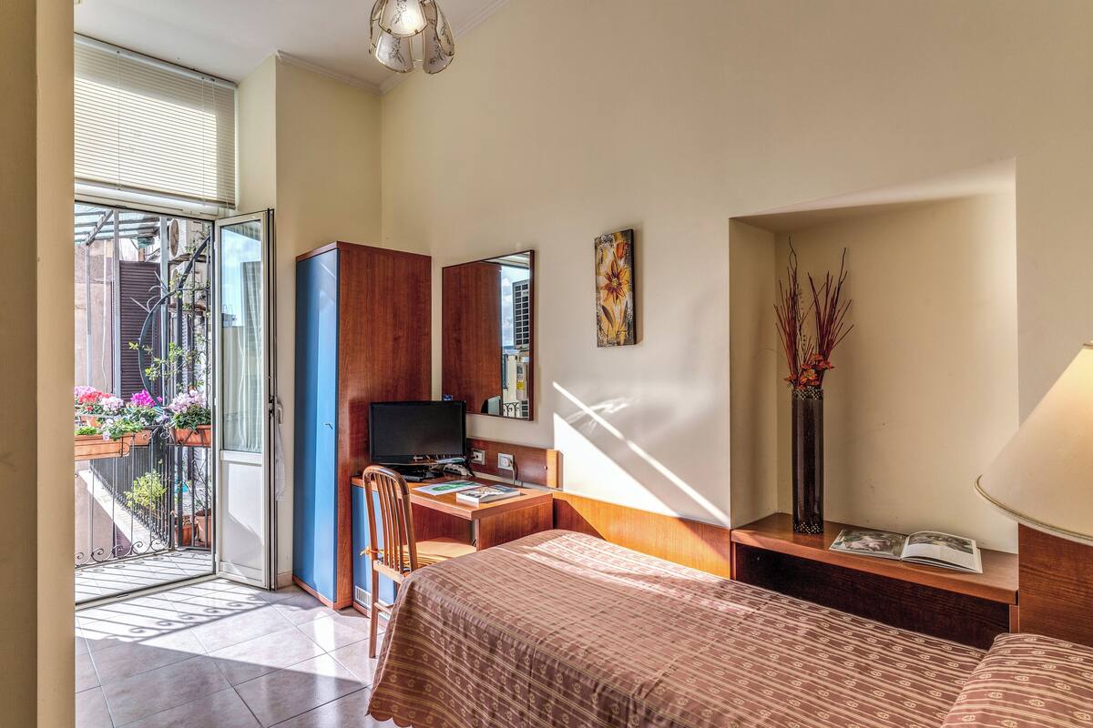 A hotel room with a bed, a desk with a computer, a chair, a TV, a window with blinds, and a balcony with potted plants.
