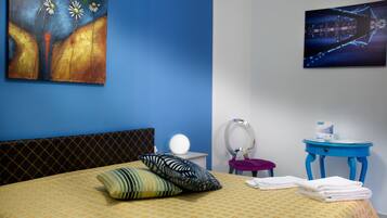 Quarto de hotel com cama, almofadas, mesa de cabeceira com abajur, espelho, pequena mesa redonda com uma xícara e obras de arte nas paredes.