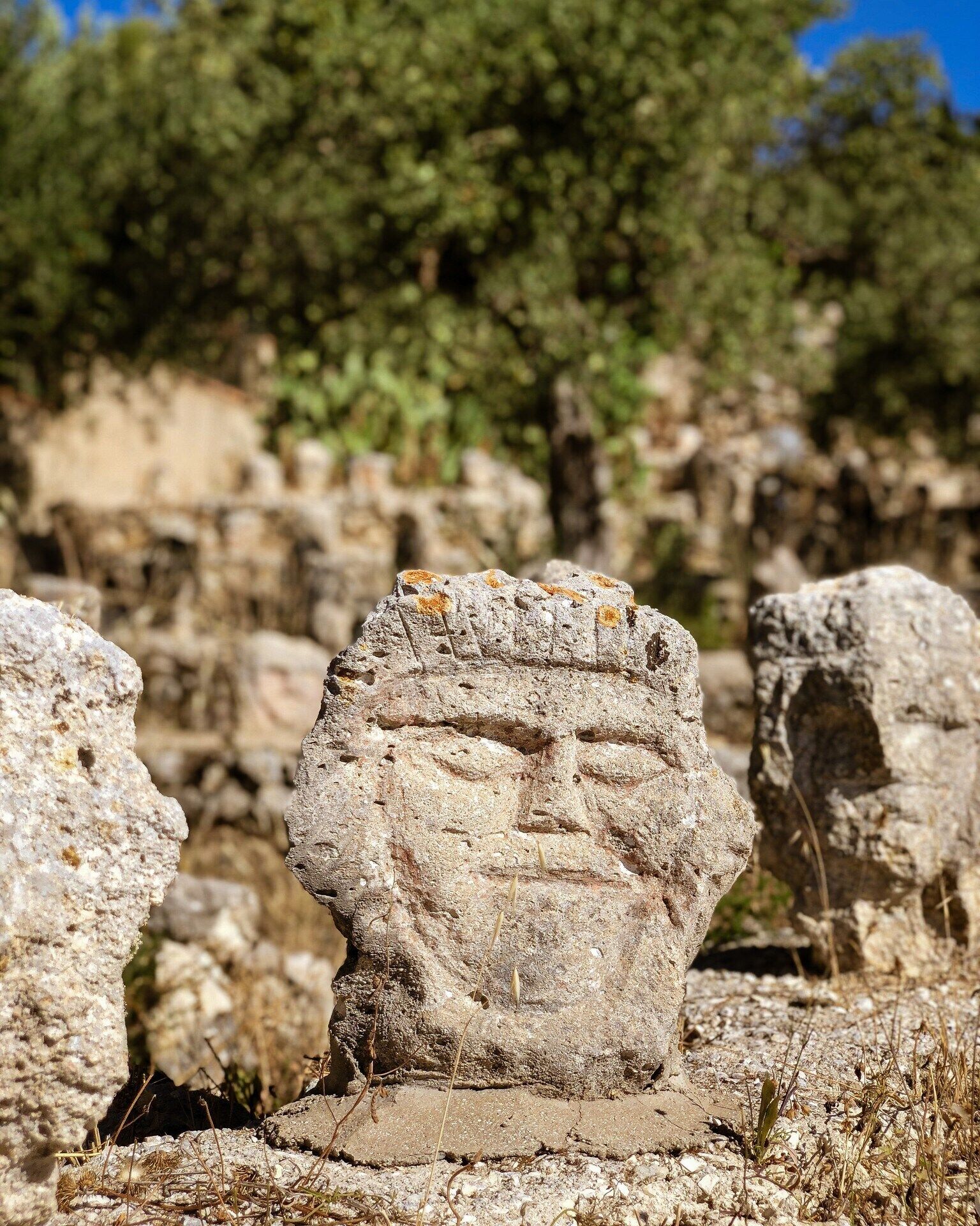 A stone structure with a carved face, surrounded by other rocks and dry vegetation.