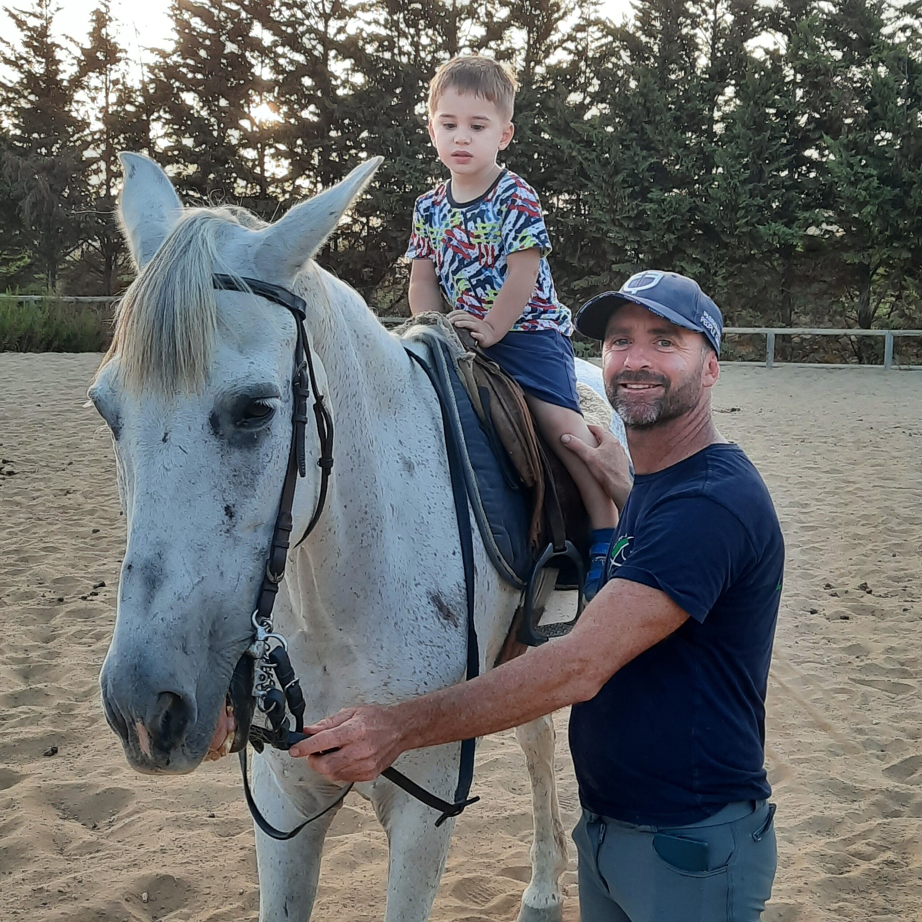 Un hombre y un niño montan un caballo blanco en una zona arenosa con árboles al fondo.
