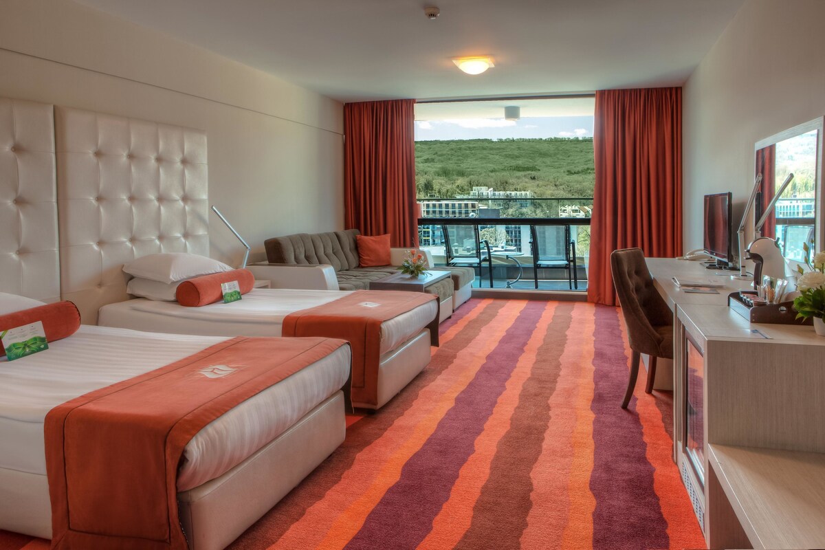Une chambre d’hôtel avec trois lits, un bureau, une chaise et un balcon donnant sur un paysage verdoyant.