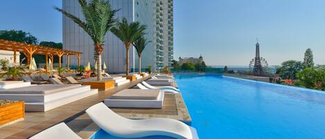 A poolside area with lounge chairs and palm trees, a tall building in the background, and a Ferris wheel.