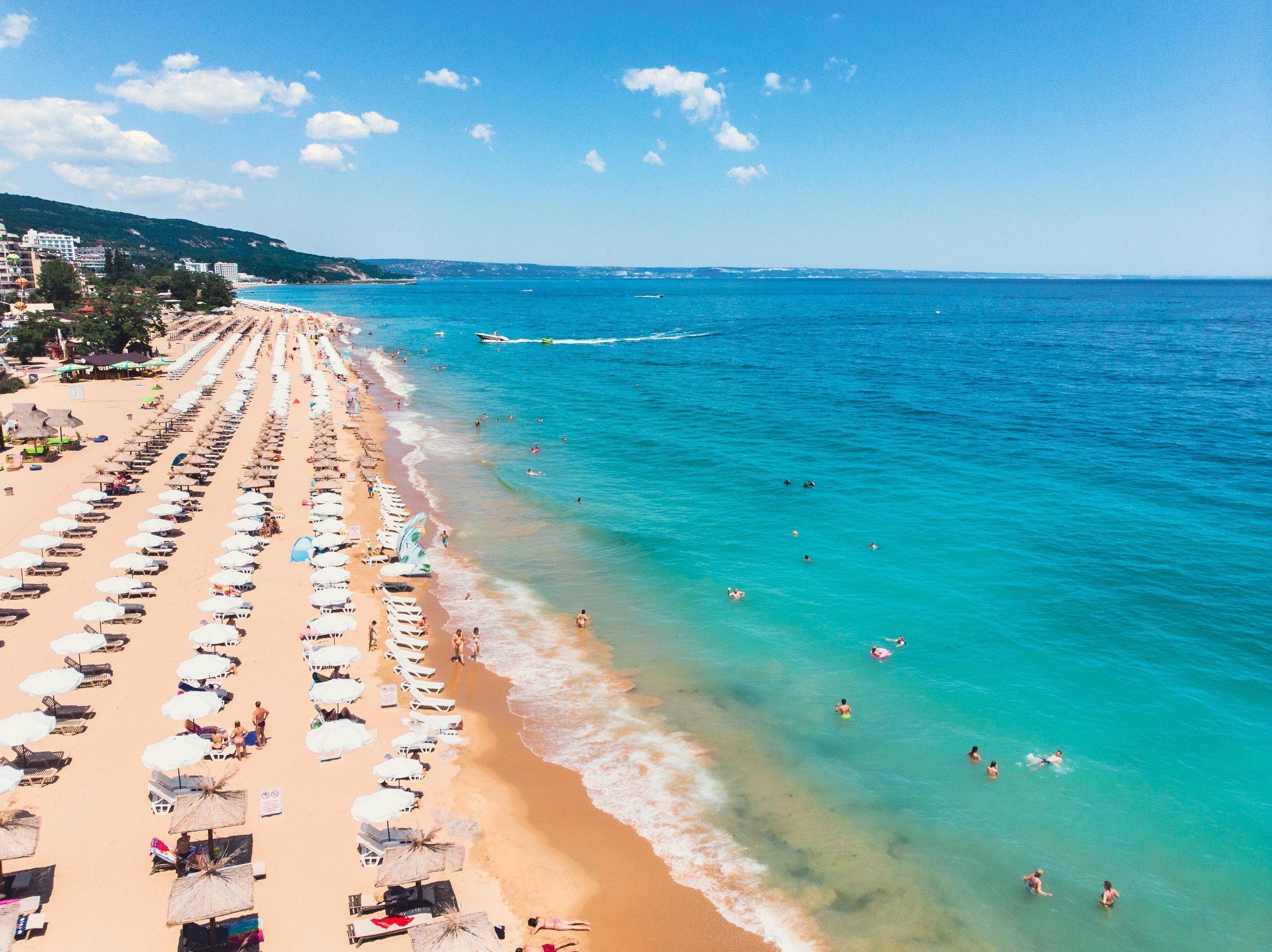 A beach with rows of sunbeds and umbrellas, a clear blue sea, and buildings in the background.