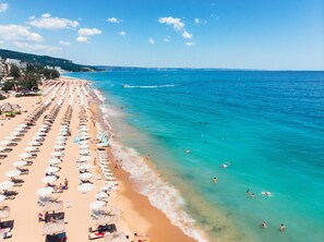 Une plage bordée de rangées de transats et de parasols, une mer d’un bleu limpide et des bâtiments en arrière-plan.