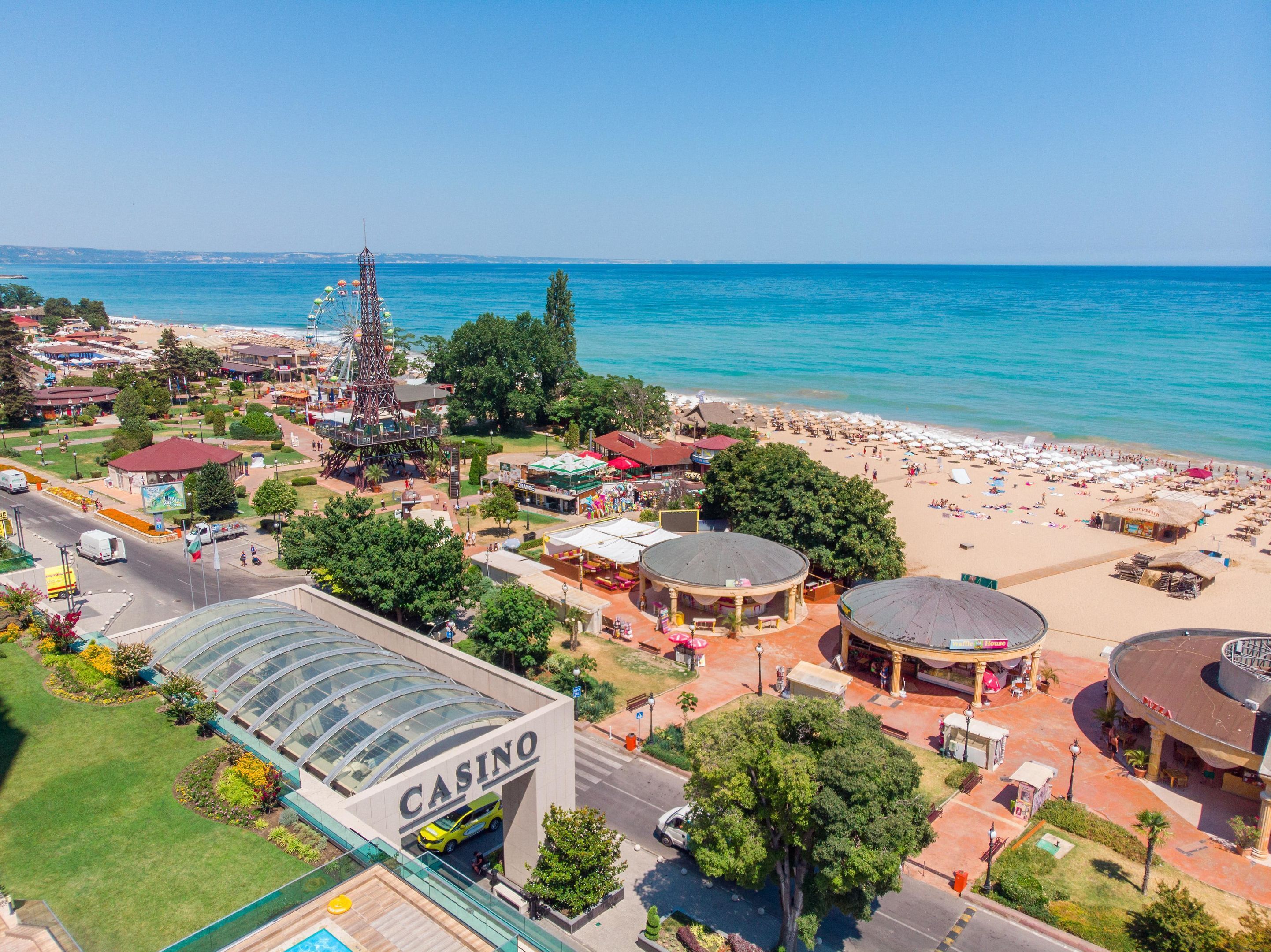 Un casino à la façade de verre, situé à côté d’une plage parsemée de parasols et où se trouve une grande roue.
