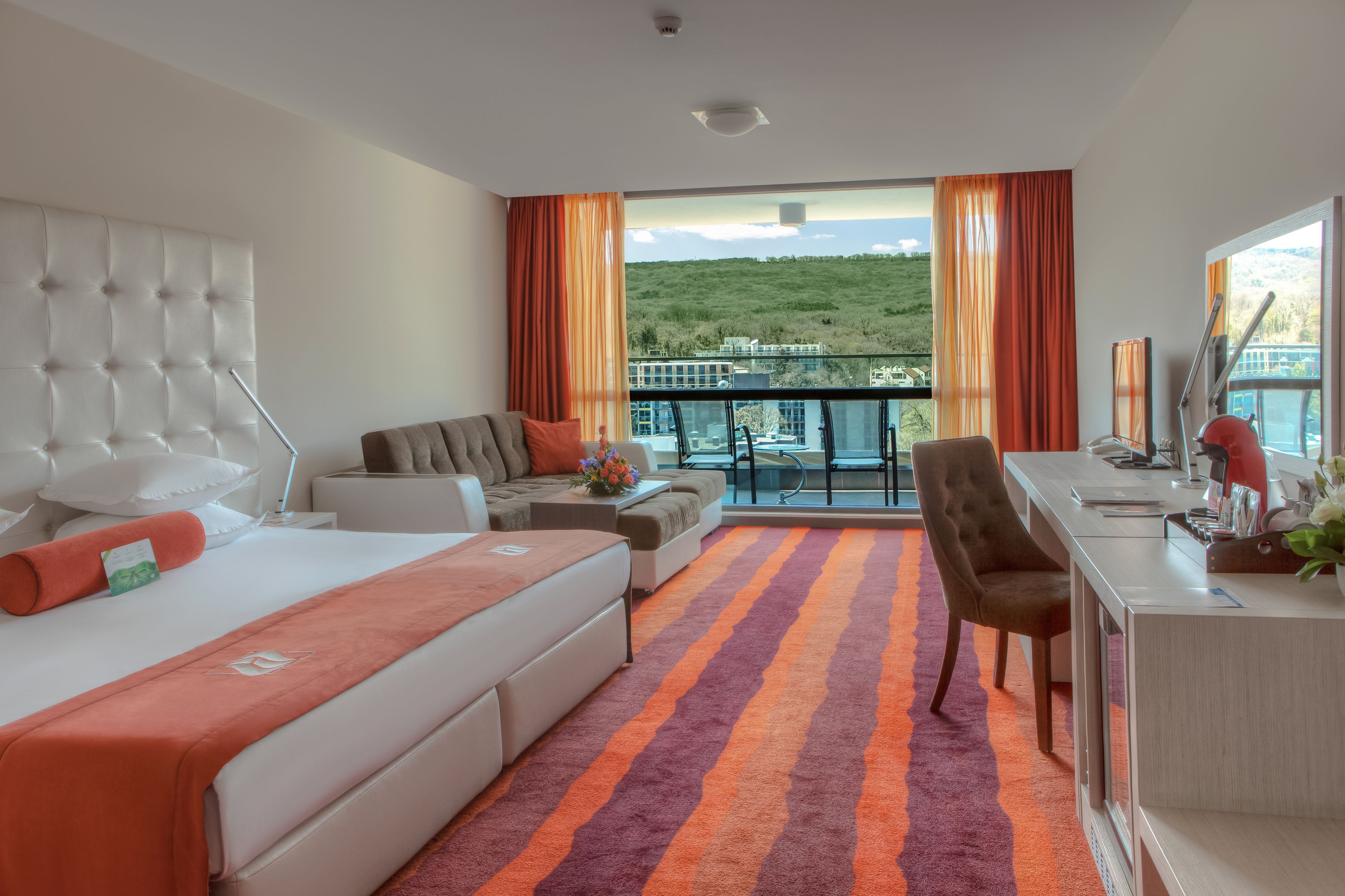 A hotel room with a bed, sofa, desk, and a balcony with a view of a hill.