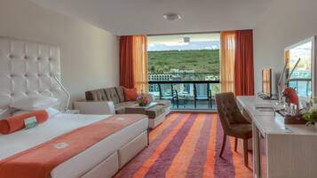 A hotel room with a bed, sofa, desk, and a balcony with a view of a hill.
