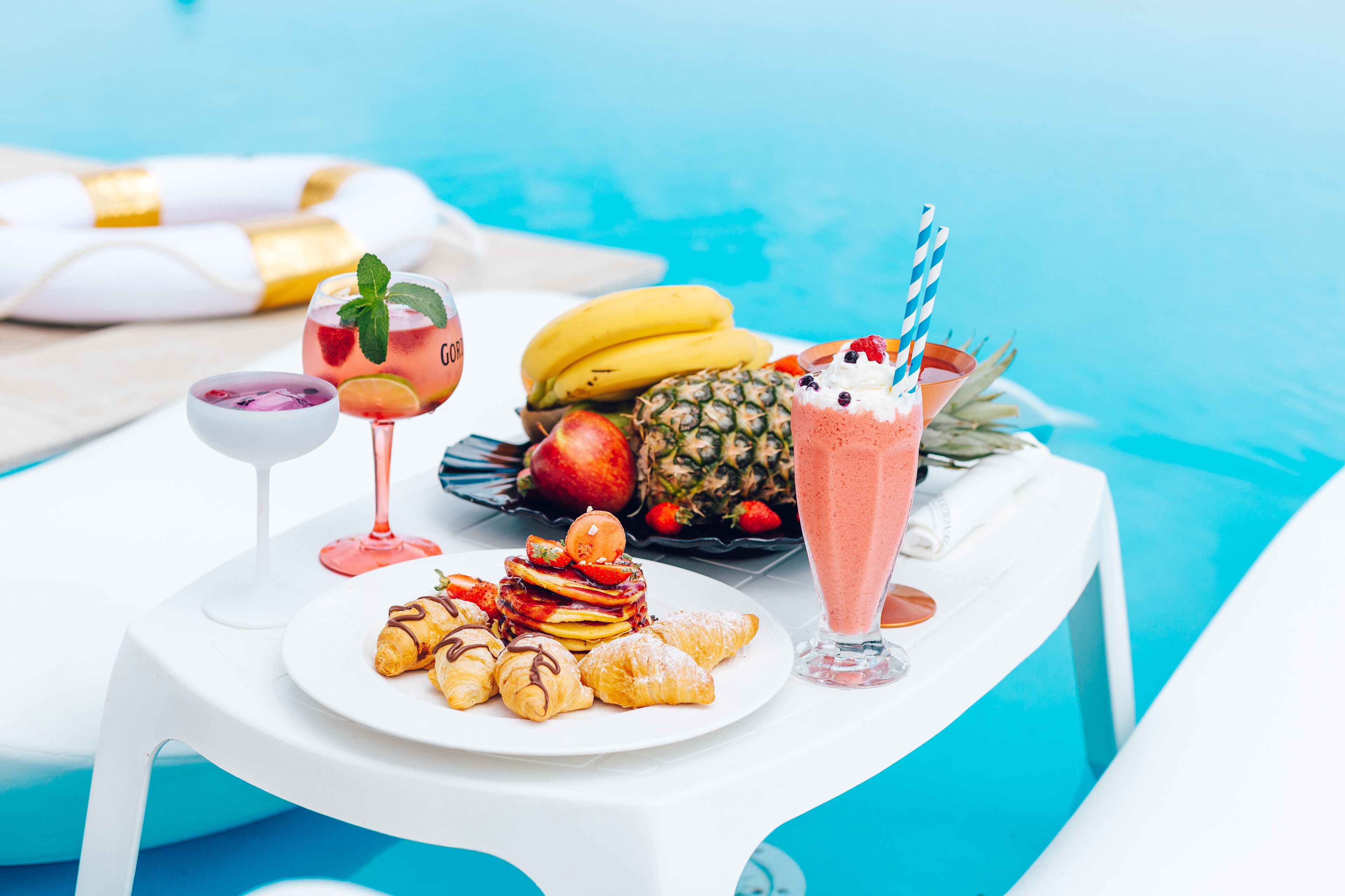 A table by a pool with a plate of pastries, a fruit salad, and two drinks.