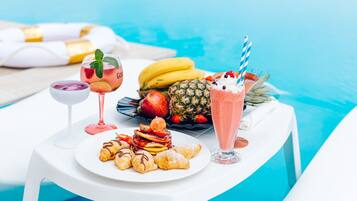 A table by a pool with a plate of pastries, a fruit salad, and two drinks.