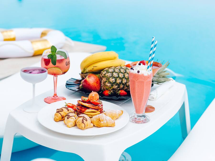 A table by a pool with a plate of pastries, a fruit salad, and two drinks.