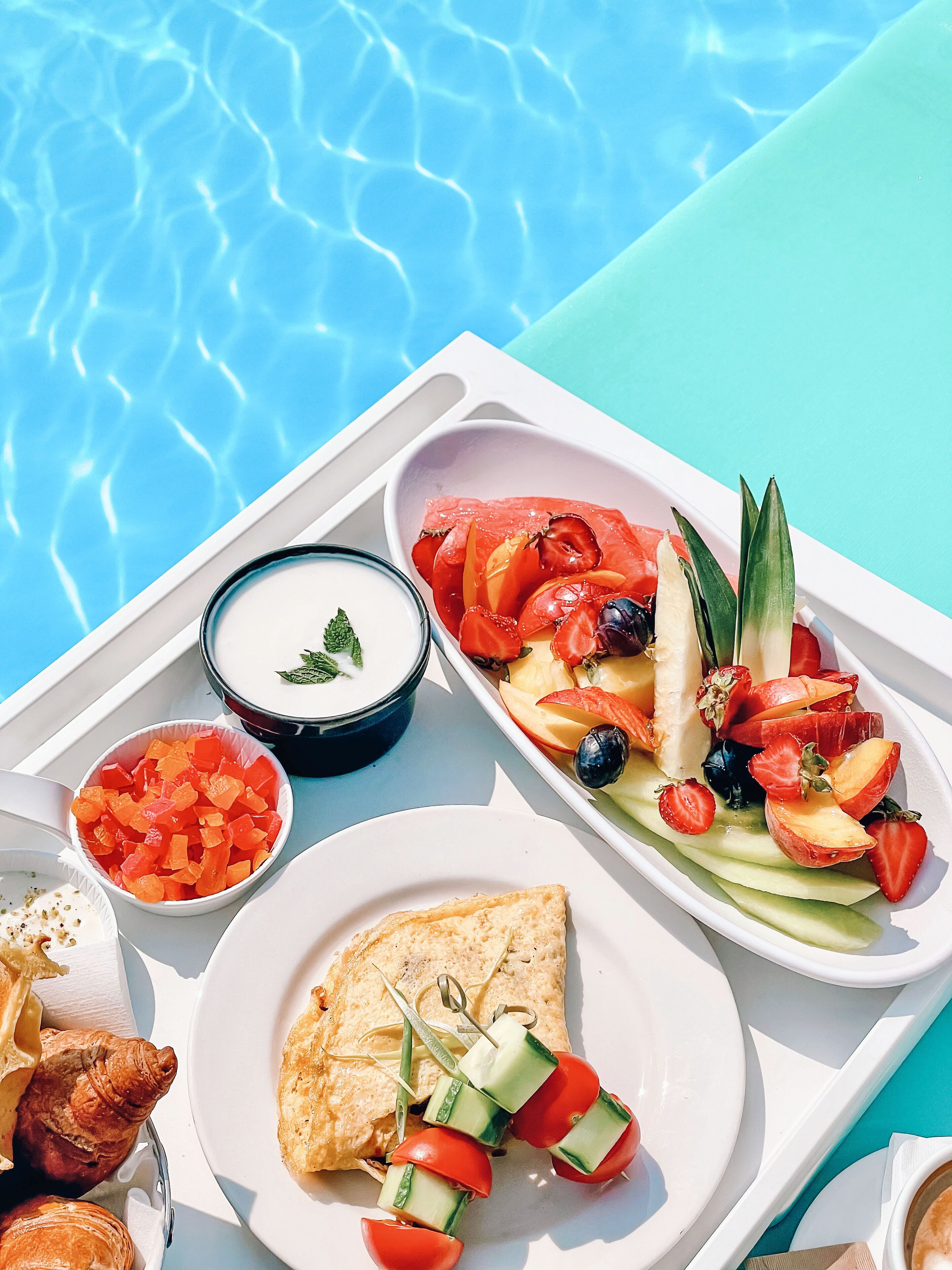 Ein Tablett mit Speisen, darunter Salat, Joghurt und Gebäck, in der Nähe eines Schwimmbeckens.