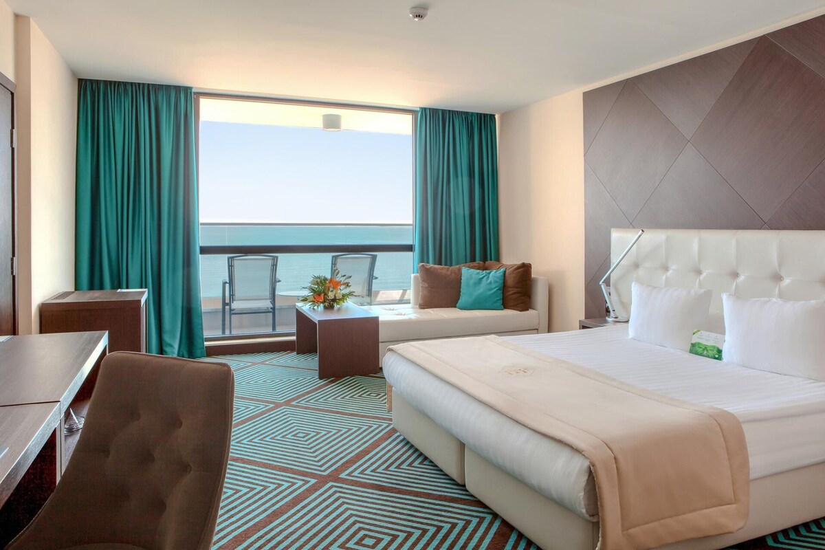 Une chambre d’hôtel avec un grand lit, un canapé, un bureau et une vue sur la mer depuis un balcon.
