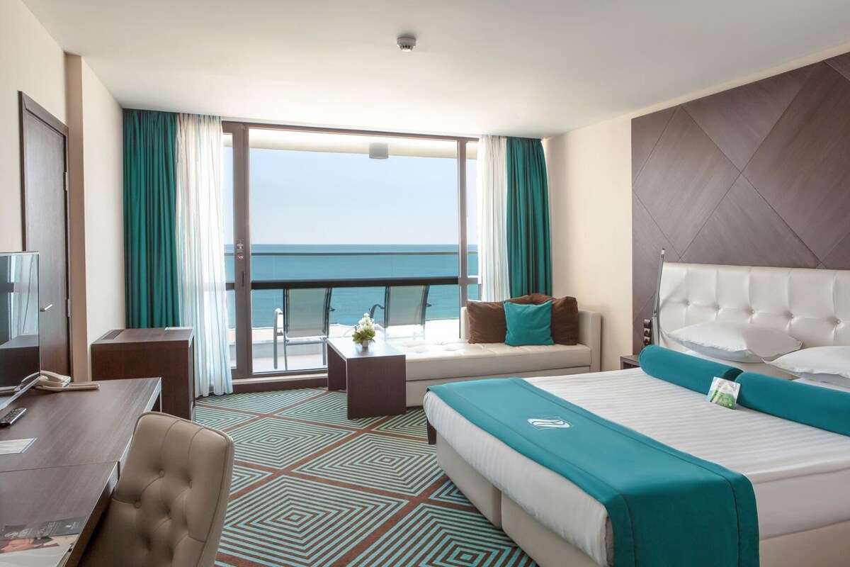 Une chambre d’hôtel avec un grand lit, un bureau, une chaise, un canapé et un balcon donnant sur la mer.
