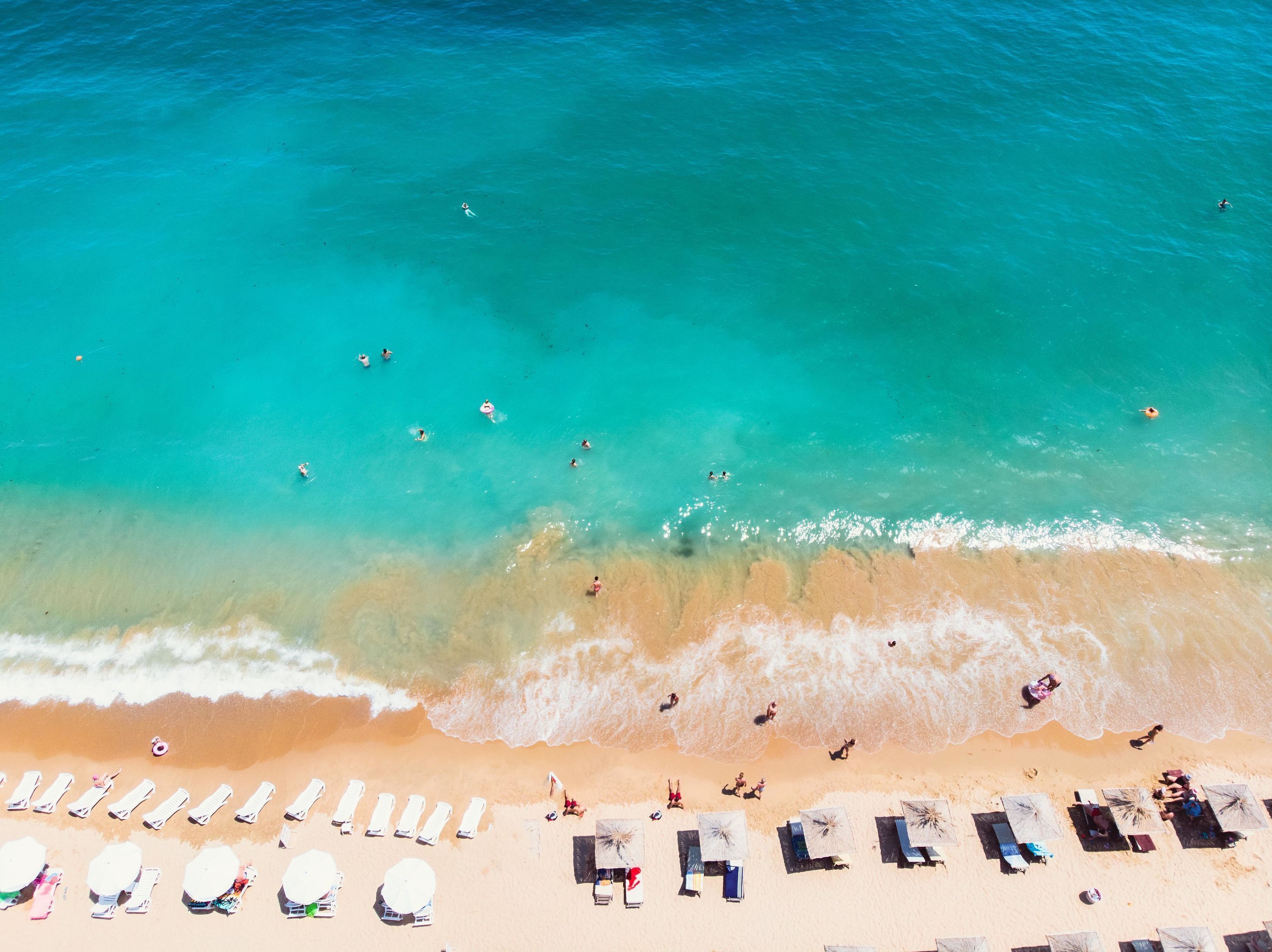 Vista aérea de una playa con aguas turquesas cristalinas, una orilla arenosa y varios sillones de baño y sombrillas.