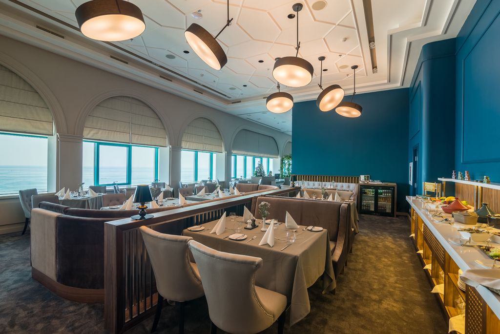 A restaurant with a long counter, multiple tables, and chairs. There are large windows with a view of the sea.