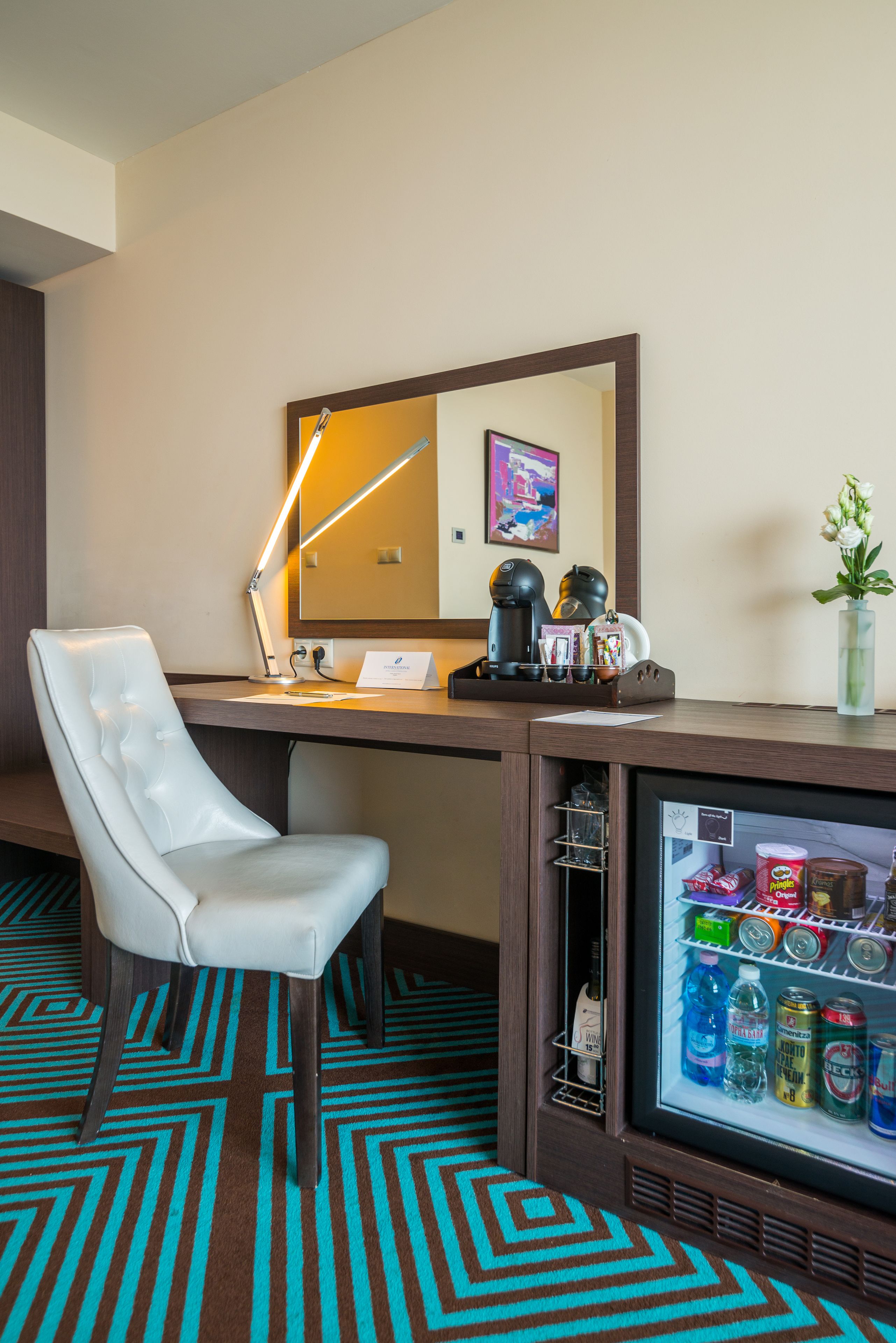 A hotel room with a white leather chair, a desk with a lamp, a refrigerator stocked with beverages, and a mirror on the wall.