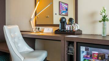 A hotel room with a white leather chair, a desk with a lamp, a refrigerator stocked with beverages, and a mirror on the wall.