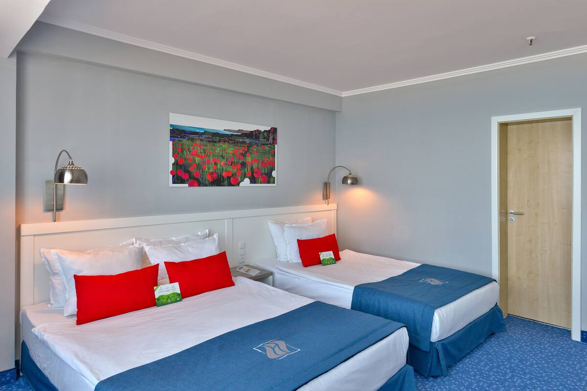 Habitación de hotel con dos camas, cada una con ropa blanca y almohadas rojas, un cuadro en la pared y una puerta de madera.