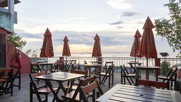 An outdoor seating area with wooden tables and chairs, orange umbrellas, and a view of the sea. - Hotel Villa Santo Antonio (Salvador)