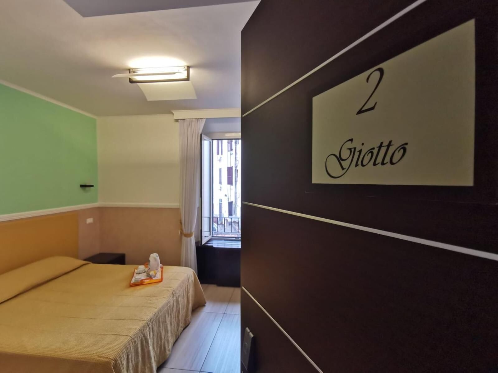 Et hotelværelse med en seng, et natbord, et lille bord med en telefon og et vindue med gardiner. Værelset har en grøn væg, et hvidt loft og en mørkebrun væg med et skilt, der står "2 Giotto".