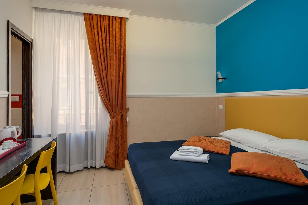 A hotel room with a bed, a desk with a chair, and a window with curtains.