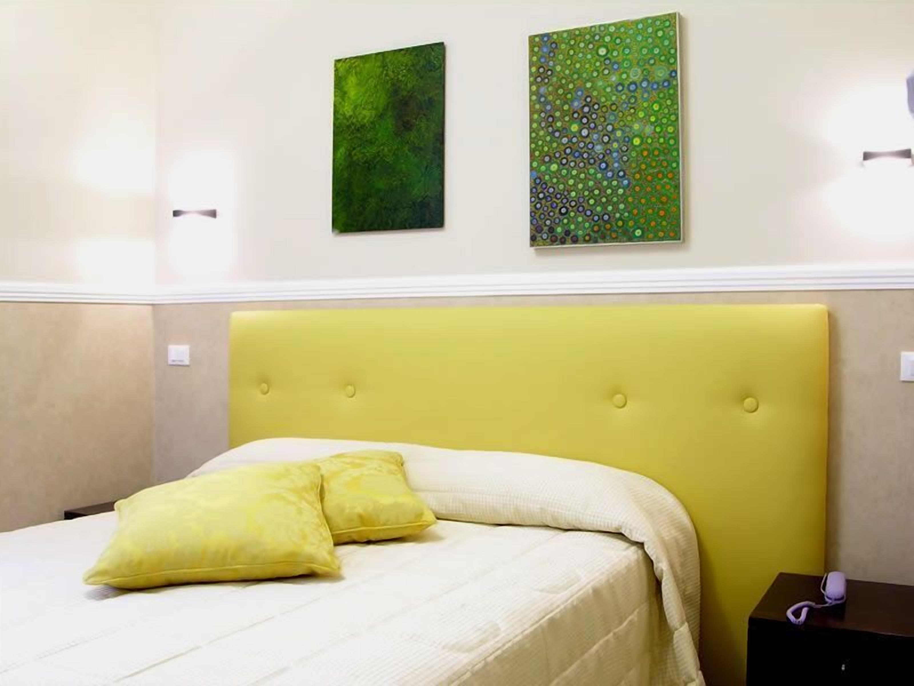 A bed with a yellow headboard, white bedding, and two yellow pillows. There are two artworks on the wall above the bed.