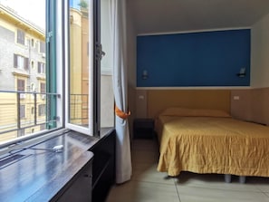 A hotel room with a bed, a window with curtains, a blue wall, and a view of buildings outside. - Mocenigo Vatican Suites (Rome)