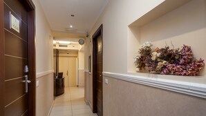 A hotel hallway with a wooden door on the left, a clock on the wall, and a floral arrangement on the right. - Mocenigo Vatican Suites (Rome)