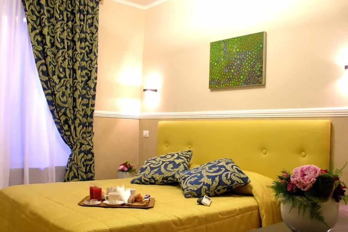 A hotel room with a yellow bedspread, decorative pillows, a tray with breakfast items, and a vase with flowers.