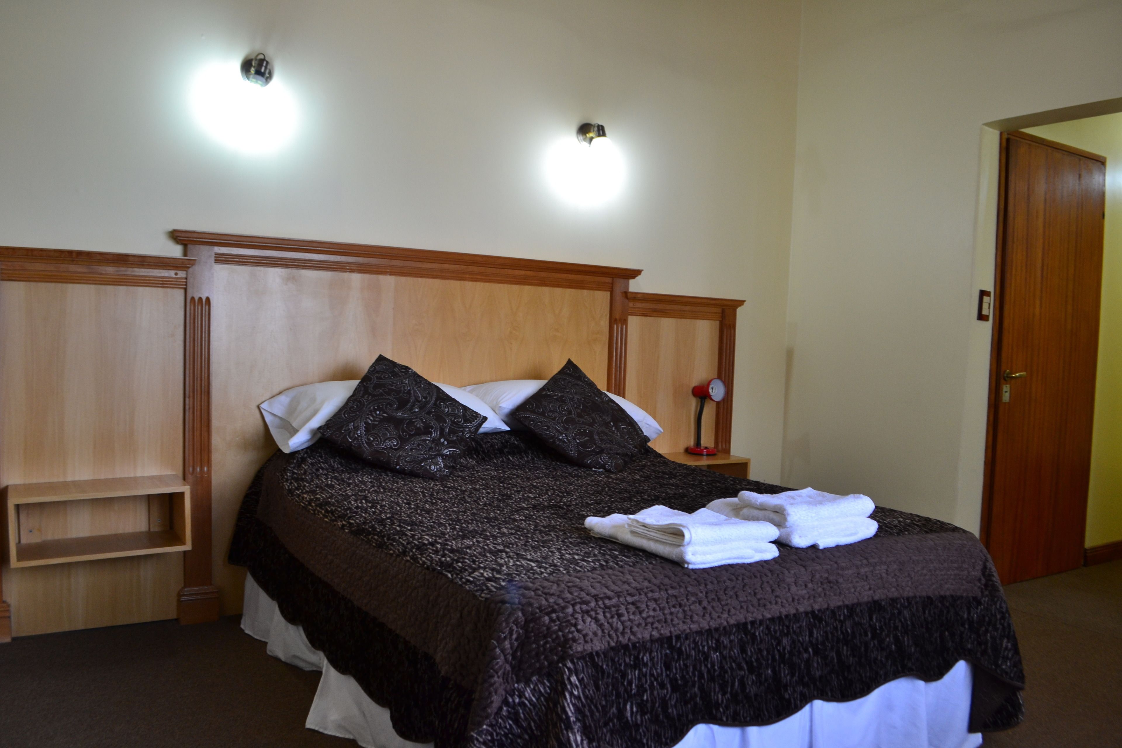 A neatly made bed with a dark blanket and pillows, a bedside table with a lamp, and a wooden headboard.