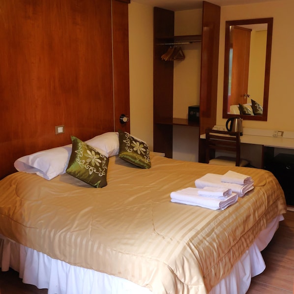 A hotel room with a large bed, a wooden wardrobe, a mirror, and a small table with a kettle.