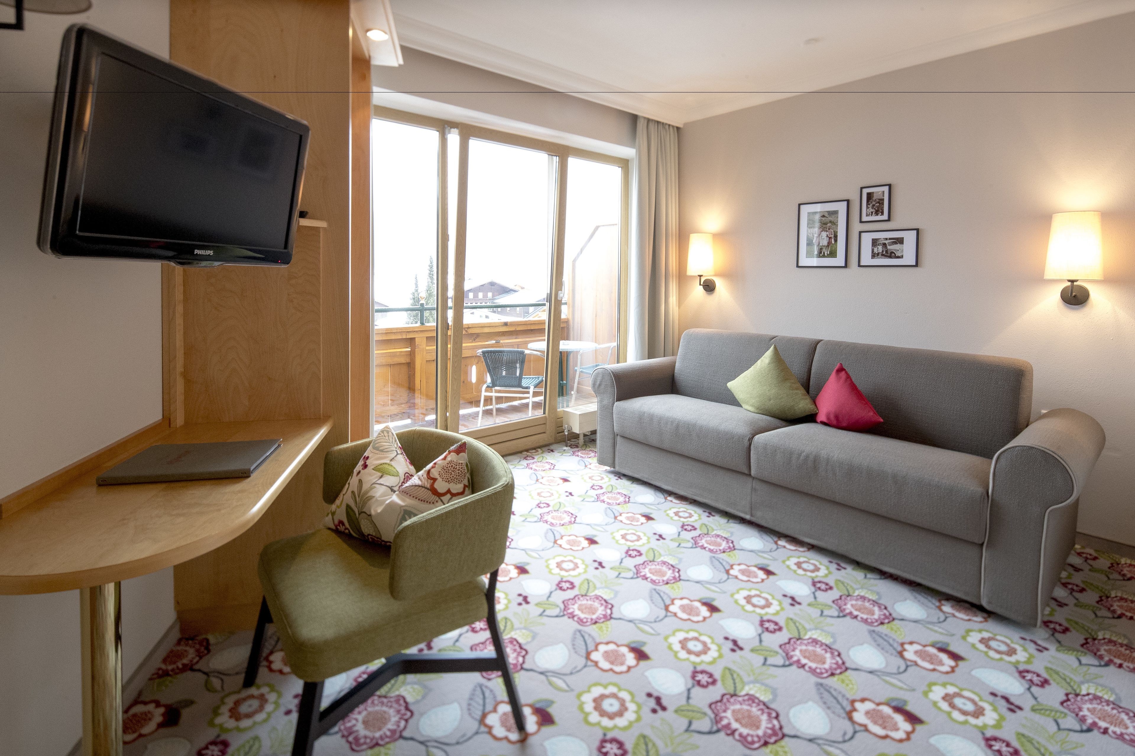 Quarto de hotel com televisão de tela plana, um sofá com almofadas coloridas, uma mesa com um laptop e um tapete estampado.