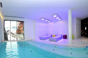 A modern indoor pool area with a clear view of the mountains through large windows, a pool with a tiled floor, and several seating areas with cushions. - Hotel Felsenhof (Flachau)