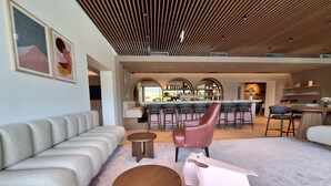 A modern living room with a sofa, a pink chair, and a small table. There is a bar area in the background with stools and a counter. - Hotel Felsenhof (Flachau)