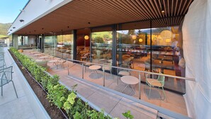 An outdoor seating area with tables and chairs, a garden bed with plants, and a modern building with large windows. - Hotel Felsenhof (Flachau)