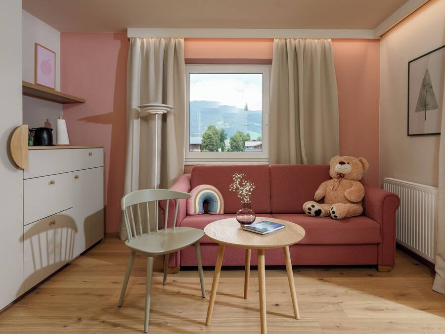 Una habitación acogedora con un sofá rosa, un osito de peluche, una mesita y una ventana con cortinas.