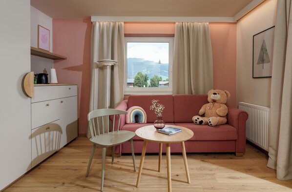 A cozy room with a pink sofa, a teddy bear, a small table, and a window with curtains. - Hotel Felsenhof (Flachau)
