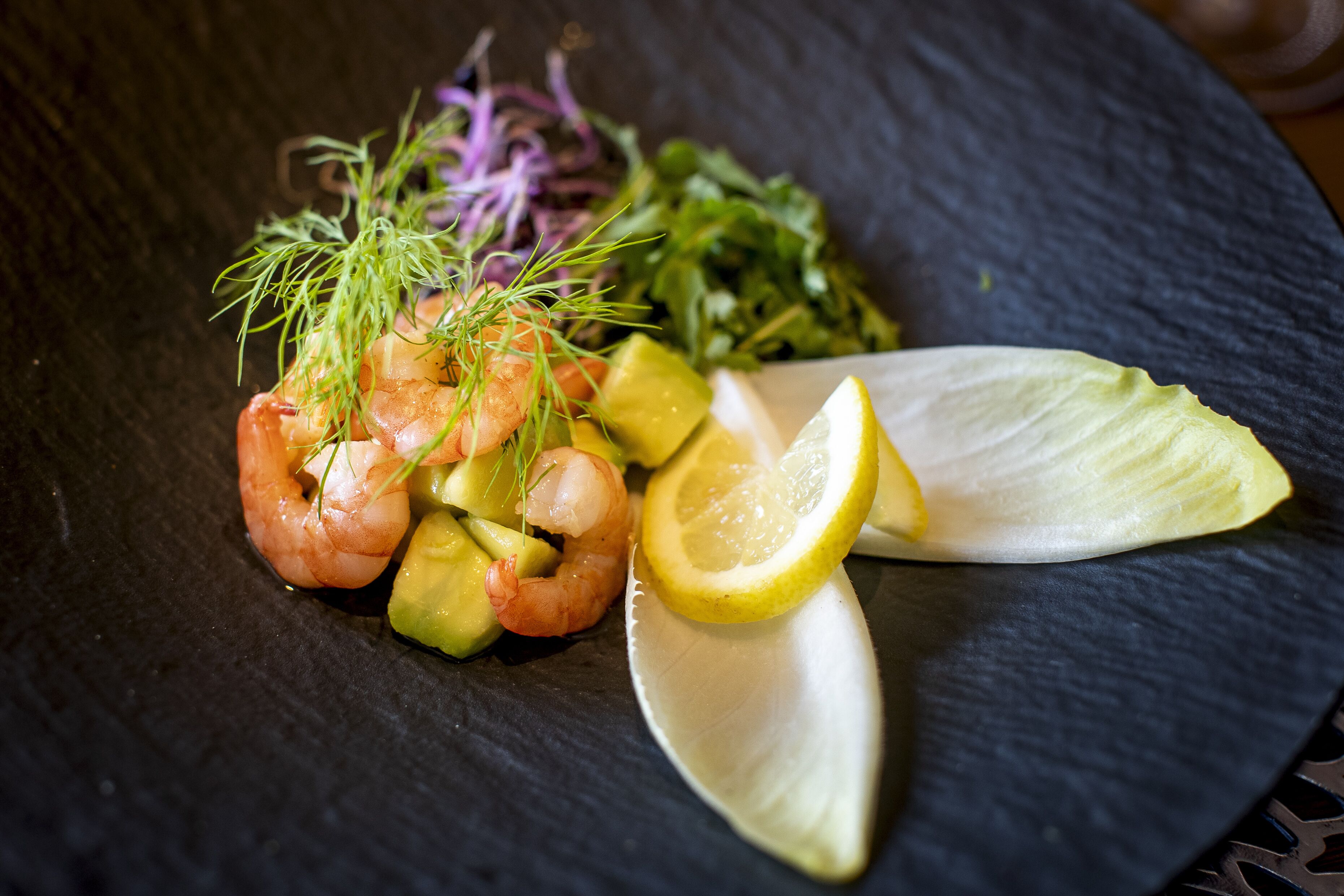 A dish with shrimp, avocado, lemon, and greens on a dark plate.