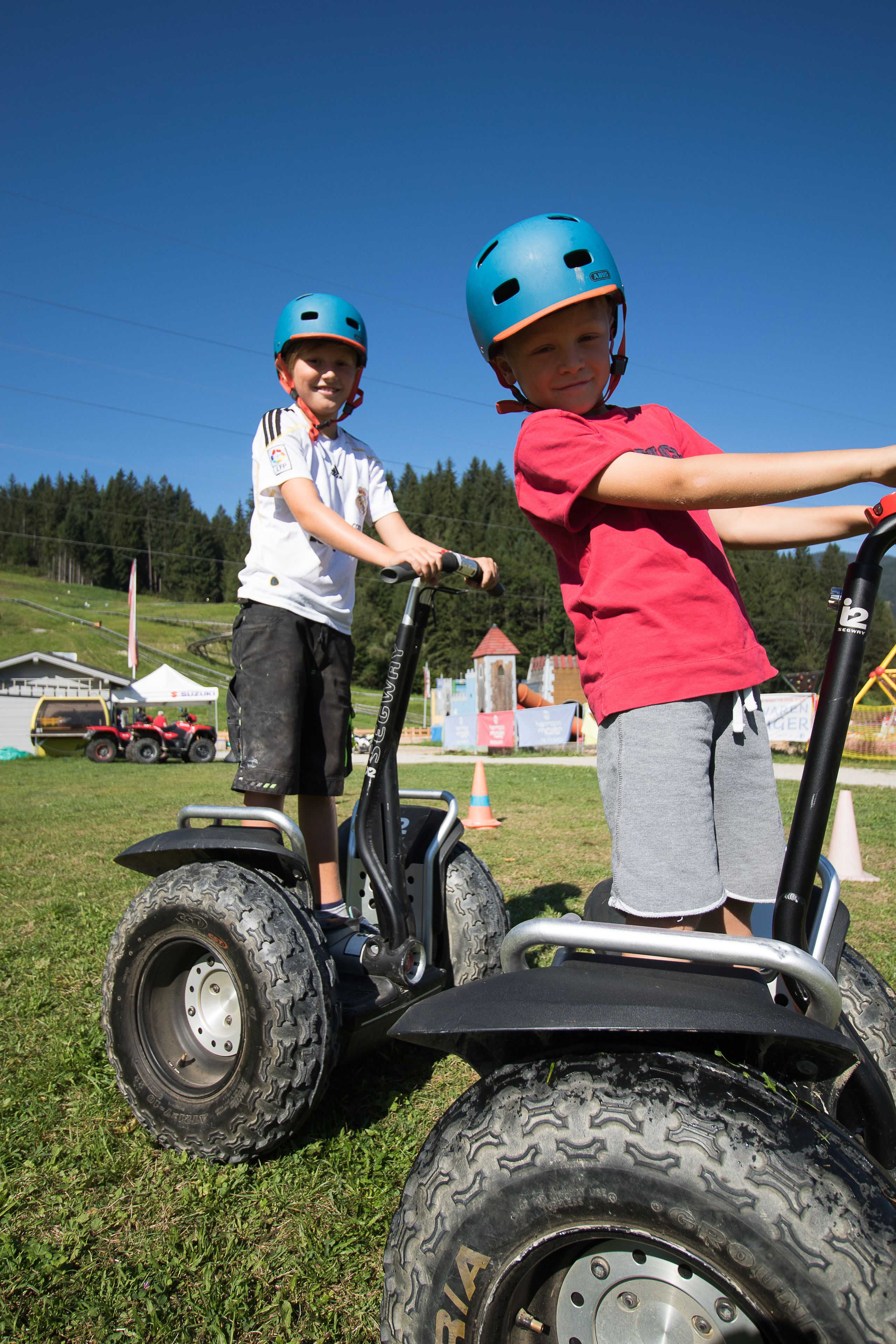 Two children on a small, four-wheeled vehicle with large tires, wearing helmets and casual clothing.