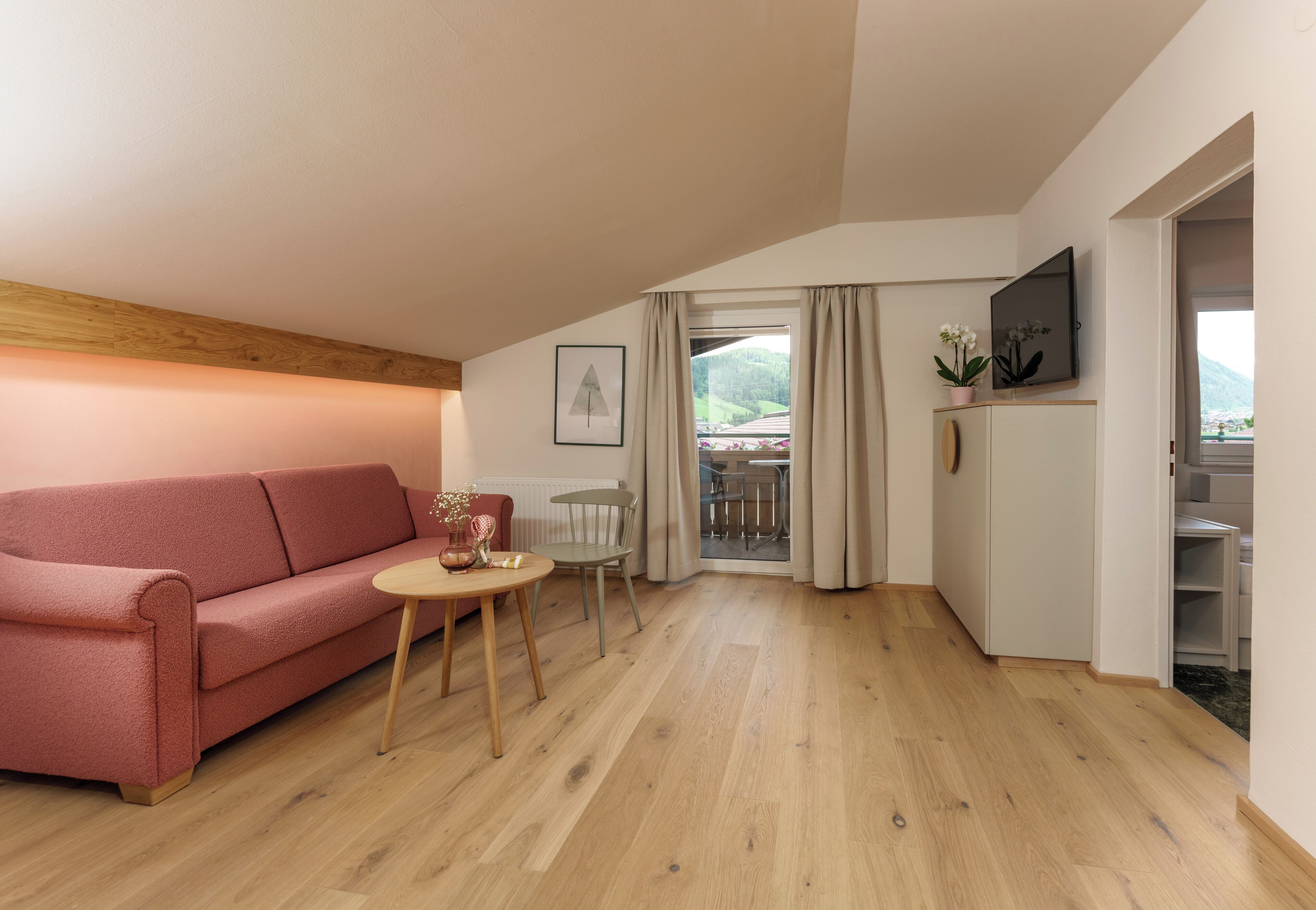 Una sala de estar acogedora con un sofá rosa, una mesita redonda y un televisor sobre un mueble. Hay un balcón con vistas a la vegetación.